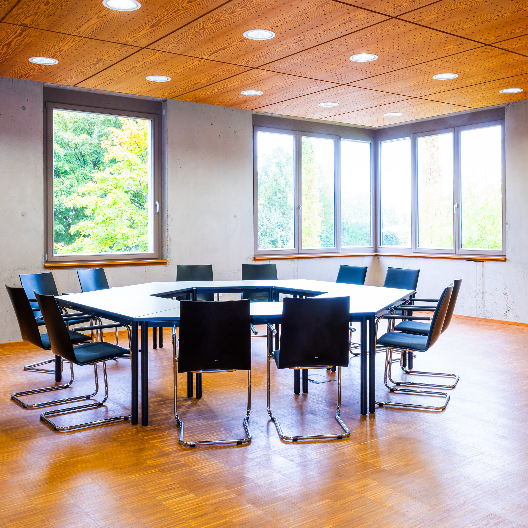 Ein heller Raum, Boden und Decke aus Holz, Wände aus Beton. Große Fenster mit Holzfensterbänken über Eck.