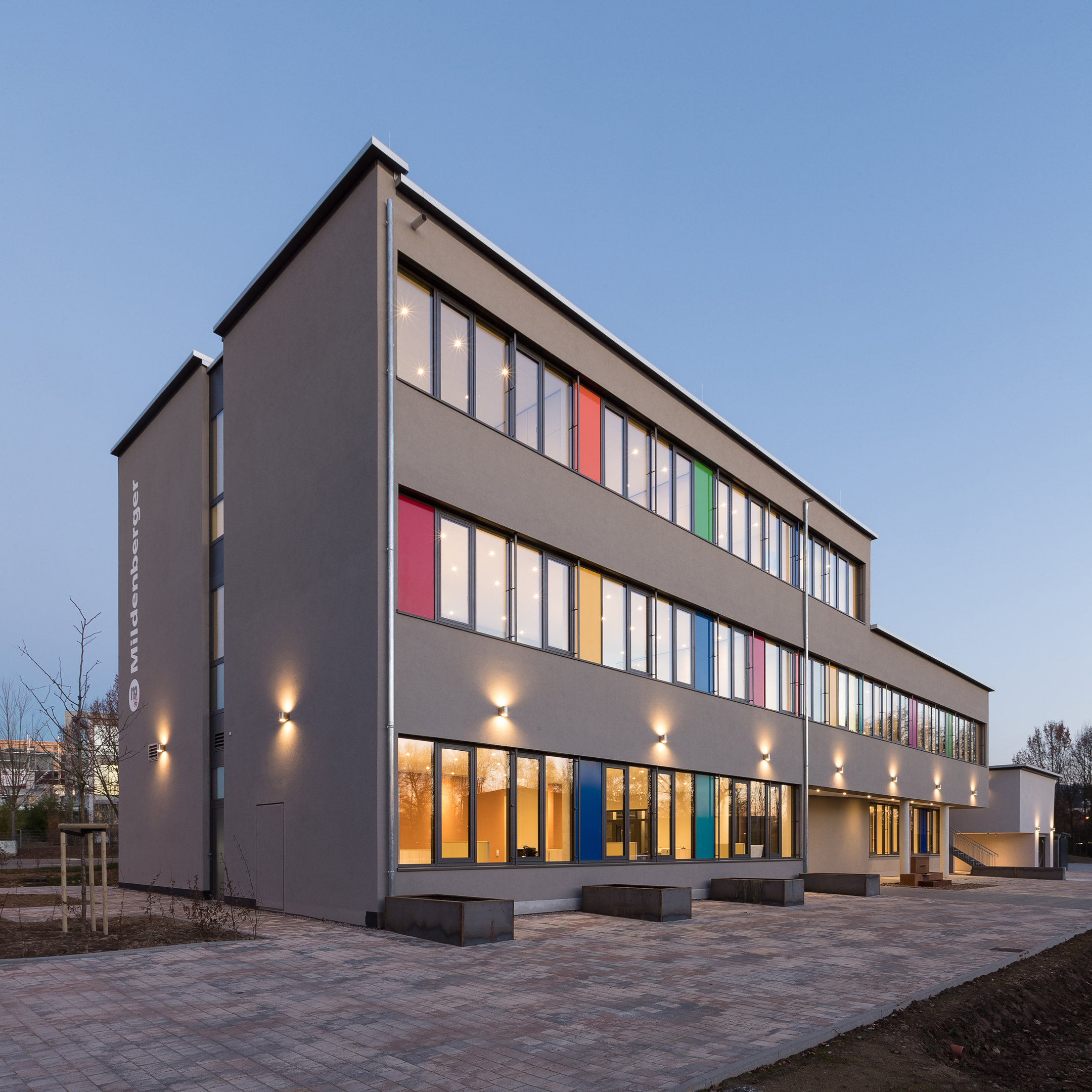 Dreistöckiges, riegelförmiges Gebäude mit Flachdach, Fassade graubeige. Die Stirnseite wird durch ein schmales, vertikales Fensterband geteilt.
