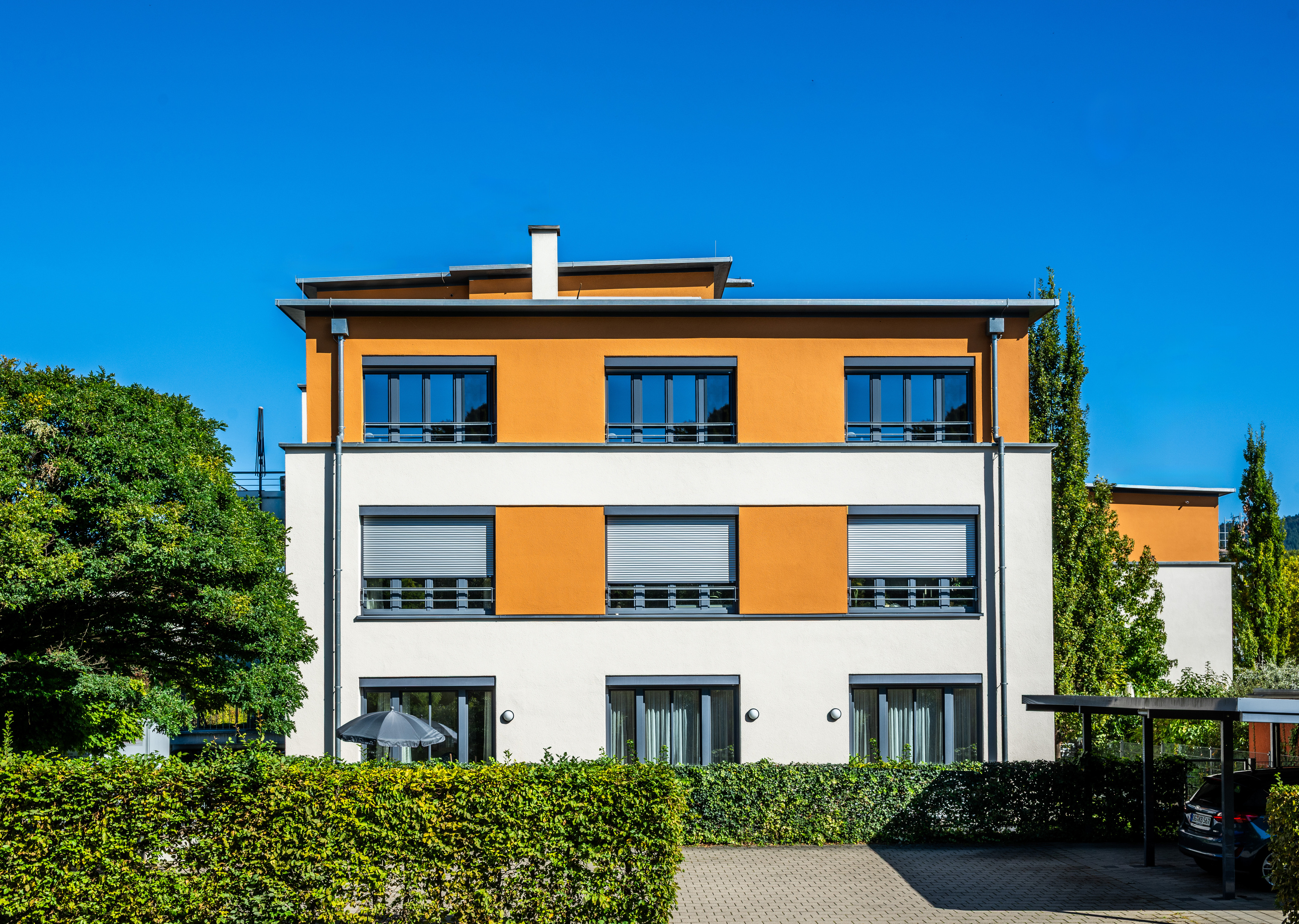 Dreistöckiger Wohnteil mit klarer Formensprache. Das Gebäude ist inmitten von Bäumen und Hecken eingegliedert.