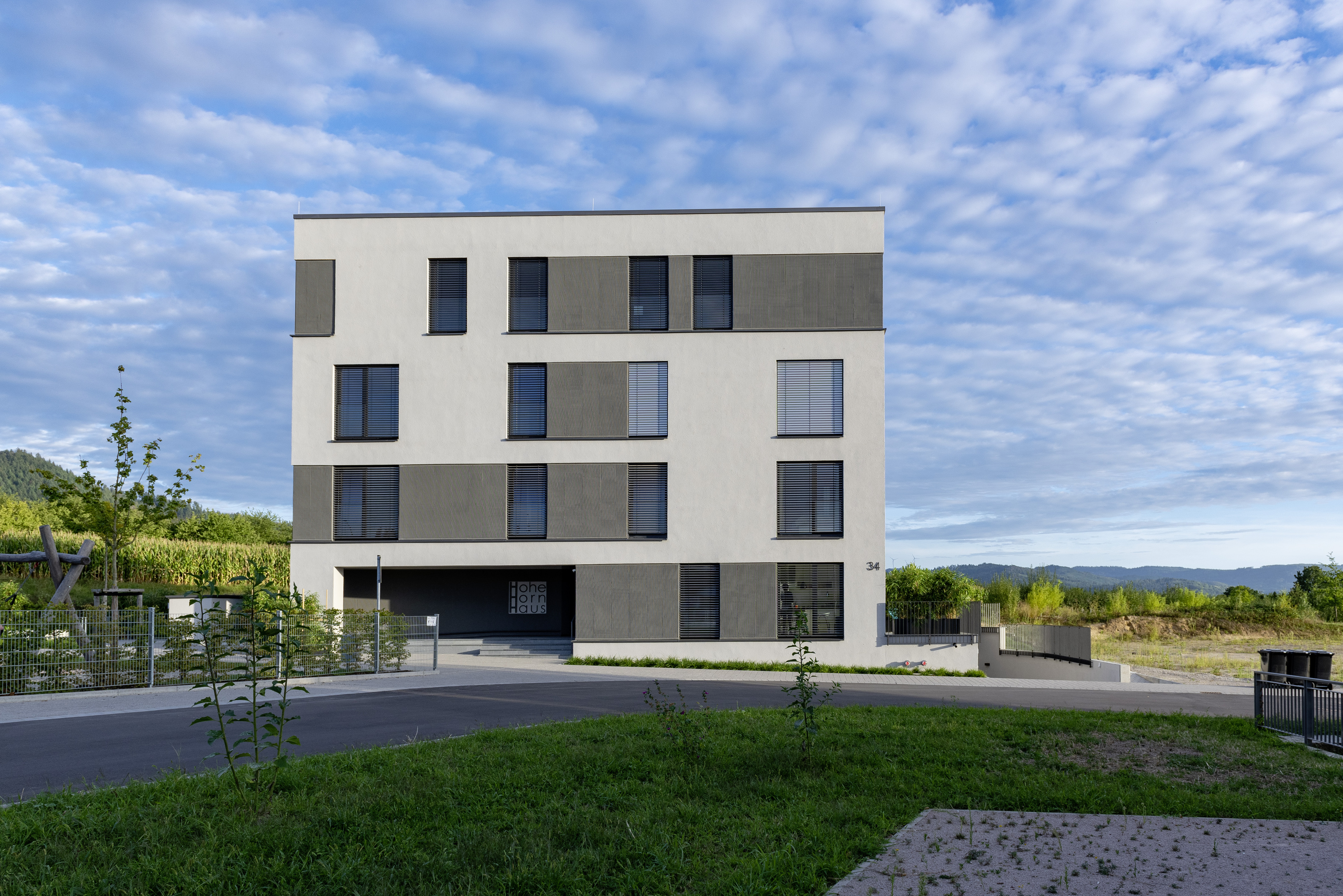 Haus mit Flachdach, frontal fotografiert. Weiße Fassade mit teils unterbrochenen dunkelgrauen Bändern um die bodentiefen Fenster.