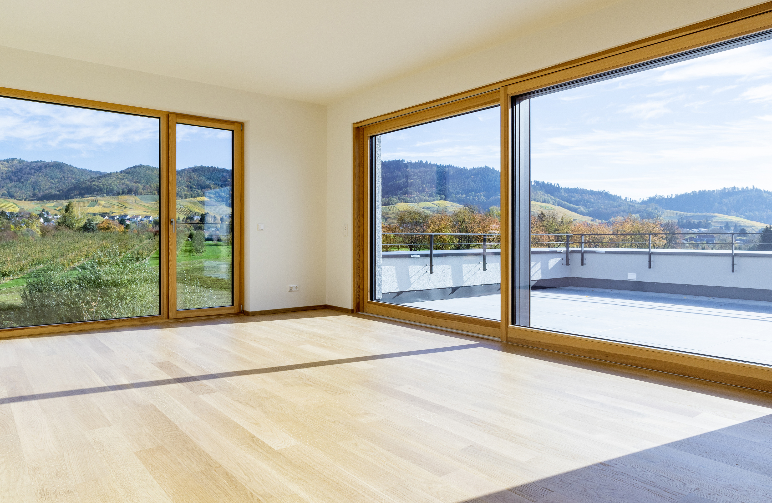 Ein weißer Raum mit Holzboden und großen, bodentiefen Fenstern. Hinterm linken Fenster eine hügelige Landschaft, hinter dem rechten eine Dachterrasse.